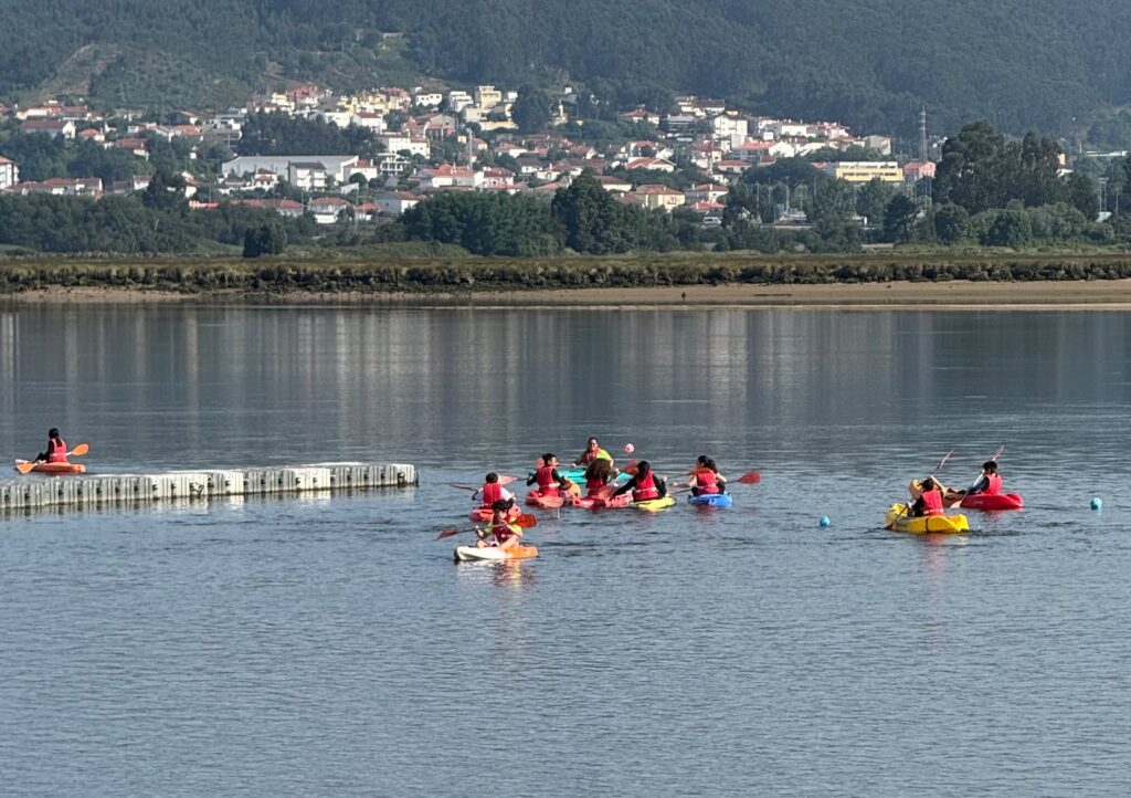 Grupo de alunos com coletes de salvação em caiaques no rio, com casario ao fundo