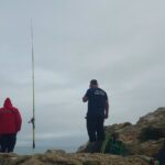 Um bombeiro e um elemento da Polícia Marítima numa zona de rochas junto ao mar, ao lado de uma cana de pesca