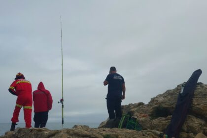 Um bombeiro e um elemento da Polícia Marítima numa zona de rochas junto ao mar, ao lado de uma cana de pesca
