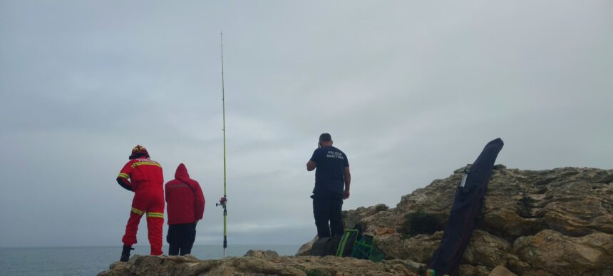 Um bombeiro e um elemento da Polícia Marítima numa zona de rochas junto ao mar, ao lado de uma cana de pesca